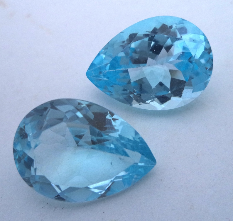 May include: Two teardrop-shaped, faceted, light blue gemstones. The gemstones are clear and have a slight sparkle.