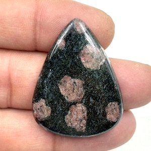 Op de afbeelding: Een zwarte en roze edelsteen cabochon met een traanvorm. De edelsteen heeft een gevlekt patroon met roze en zwarte vlekken.