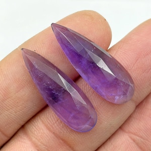 Puede incluir: Dos piedras preciosas de amatista facetadas en forma de lágrima. Las piedras preciosas son de color púrpura intenso con una punta. Las facetas capturan la luz, creando un efecto brillante. Las piedras preciosas se muestran sobre un fondo neutro.