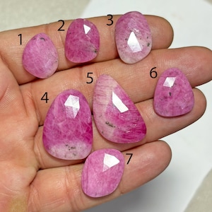 Puede incluir: Siete piedras preciosas rosas facetadas con inclusiones naturales, numeradas del uno al siete. Cada piedra presenta una forma irregular pulida con una parte superior plana y una superficie reflectante, mostrando tonos de magenta y rosa claro.