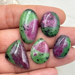 Puede incluir: Cinco piedras preciosas pulidas de varias formas, incluyendo ovaladas, en forma de lágrima y triangulares. Las piedras presentan una vibrante mezcla de colores: rojo intenso, verde y negro. Las piedras preciosas se sostienen en una mano, mostrando su belleza natural y patrones únicos.