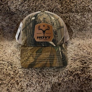May include: Camouflage baseball cap with a brown leather patch featuring a deer antler design and the text "HOYT ARCHERY". The cap has a mesh back and a curved brim.
