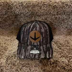 May include: A camouflage trucker hat with a brown leather patch featuring a downward pointing arrow. The hat has a black mesh back and a Richardson 112 adjustable strap.