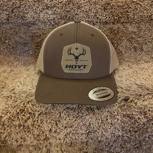 May include: A brown and tan baseball cap with a mesh back. The front of the cap features a patch with a deer skull graphic and the words "HOYT ARCHERY" and "SINCE 1931". The cap has a curved brim and a snapback closure.