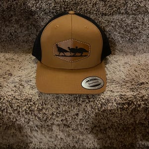 May include: A brown and black trucker hat with a snapback closure. The front panel features a leather patch with a silhouette of three turkeys. The hat has a curved brim and a mesh back.