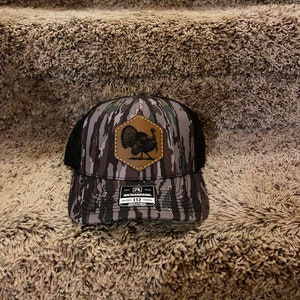 May include: A camouflage trucker hat with a brown leather patch featuring a turkey illustration. The hat has a black mesh back and a Richardson 112 adjustable strap.