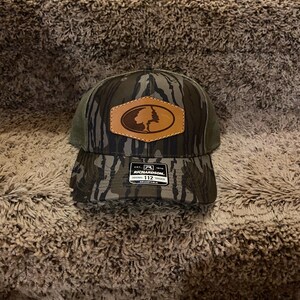 May include: Camouflage baseball cap with a brown leather patch featuring a tree logo. The cap has a green mesh back and a curved brim. The Richardson brand label is visible.