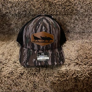May include: A camouflage-patterned trucker hat with a black mesh back. The front features a brown leather patch with silhouettes of three turkeys. The hat has a curved brim and an adjustable strap.