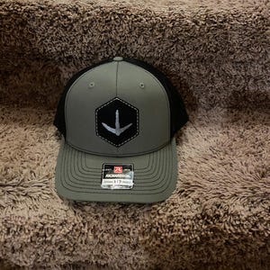 May include: A gray and black trucker hat with a structured front panel and a mesh back. The front features a patch with a white bird footprint design. The hat has a curved brim and a snapback closure. The brand name "Richardson" is visible on the brim.