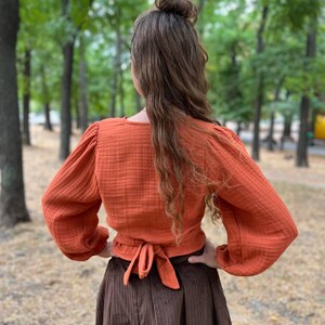 May include: An orange wrap top with long, puffed sleeves and a tie closure at the waist. The top is paired with a long, brown, pleated skirt. The outfit is made of textured fabric and is worn outdoors.
