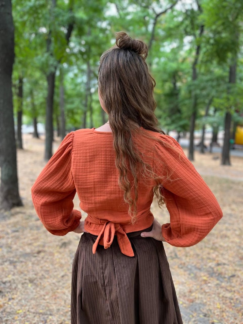 May include: An orange wrap top with long, puffed sleeves and a tie closure at the waist. The top is paired with a long, brown, pleated skirt. The outfit is made of textured fabric and is worn outdoors.