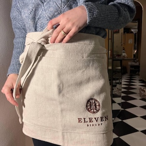 May include: A beige linen apron with an embroidered logo that says "Eleven Distillery".