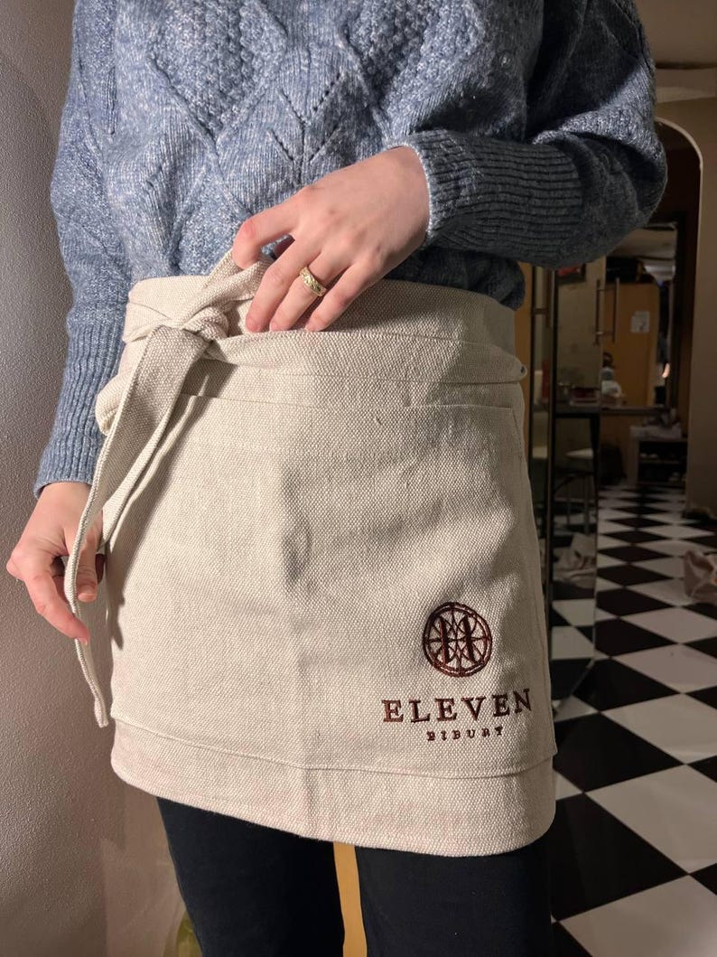 May include: A beige linen apron with an embroidered logo that says "Eleven Distillery".