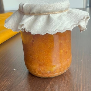 Puede incluir: Un pequeño tarro de cristal lleno de conservas de color naranja. El tarro está cubierto con una tapa de tela blanca, asegurada con hilo. El tarro se encuentra sobre una superficie de madera oscura, lo que sugiere un producto artesanal.