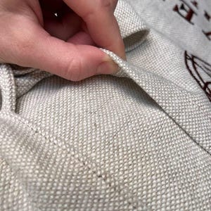 May include: Close-up of a beige canvas tote bag with the text "HE" embroidered in brown thread.