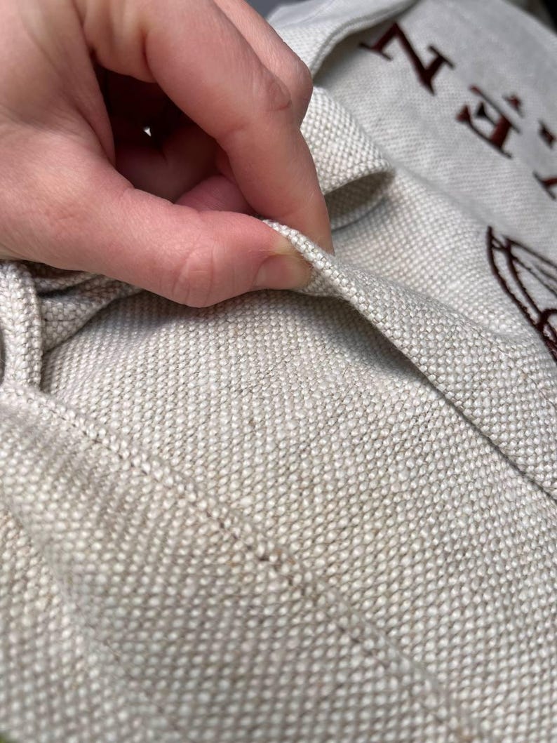 May include: Close-up of a beige canvas tote bag with the text "HE" embroidered in brown thread.