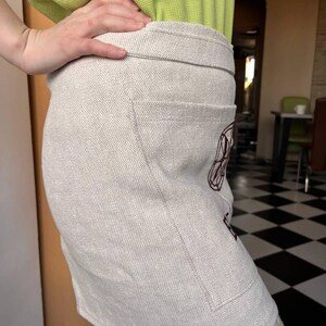 May include: A beige linen apron with a large pocket on the front. The pocket has a burgundy embroidered design.