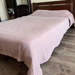 May include: A bed with a dark brown wooden frame and a light pink bedspread. The bedspread covers the mattress and pillows, with the fabric draped over the sides. The room has a wooden floor and a dark brown door.