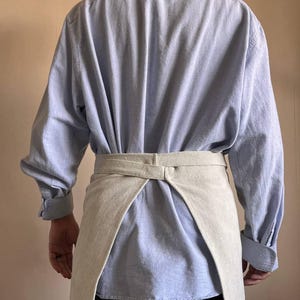 May include: A light blue long-sleeved shirt with a beige linen apron tied around the waist.