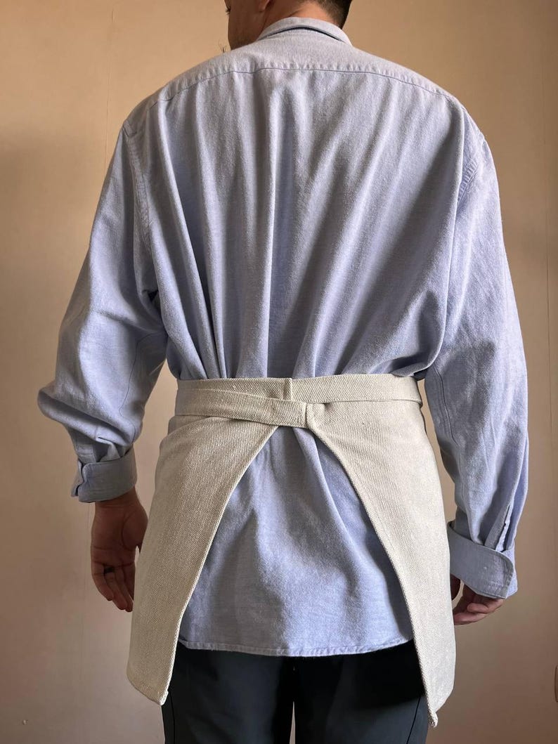 May include: A light blue long-sleeved shirt with a beige linen apron tied around the waist.