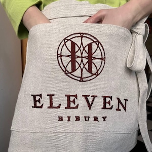 May include: A beige linen apron with a red embroidered logo featuring a circle with an "H" inside. The text "ELEVEN BIBURY" is embroidered in red below the logo.