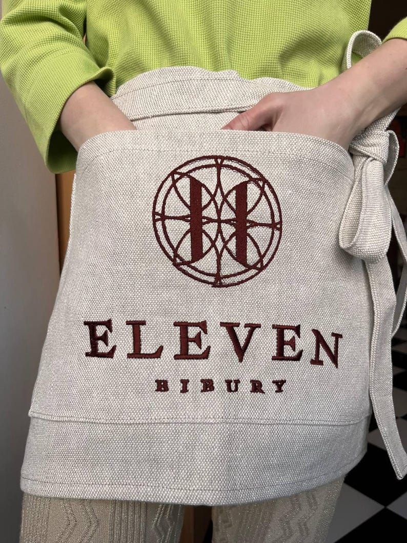 May include: A beige linen apron with a red embroidered logo featuring a circle with an "H" inside. The text "ELEVEN BIBURY" is embroidered in red below the logo.