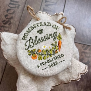 Op de afbeelding: Een ronde, beige stoffen potdeksel met de geborduurde tekst "HOMESTEAD OF Blessing ESTABLISHED 2022". De deksel heeft een bloemen- en groenteontwerp, inclusief wortelen en tomaten. De deksel is vastgebonden met een touw.