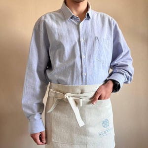 May include: A person wearing a light blue button-down shirt and a beige apron with a logo that says "Eleven Eleven Brewery".