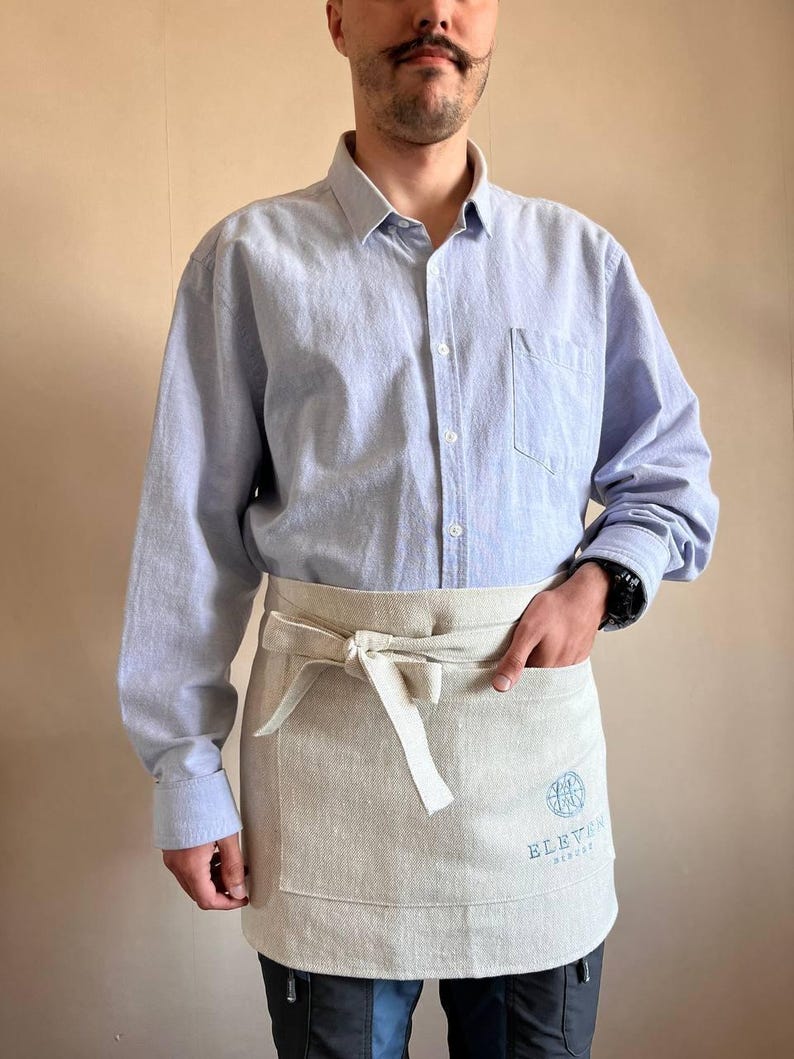May include: A person wearing a light blue button-down shirt and a beige apron with a logo that says "Eleven Eleven Brewery".