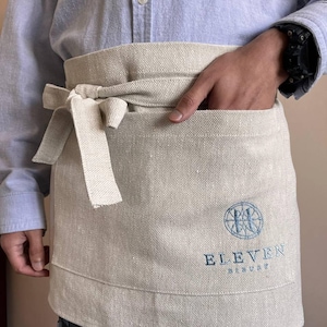May include: A beige linen apron with a large pocket and a tie. The apron has a blue embroidered logo that says "ELEVEN" and "RESTAURANT".
