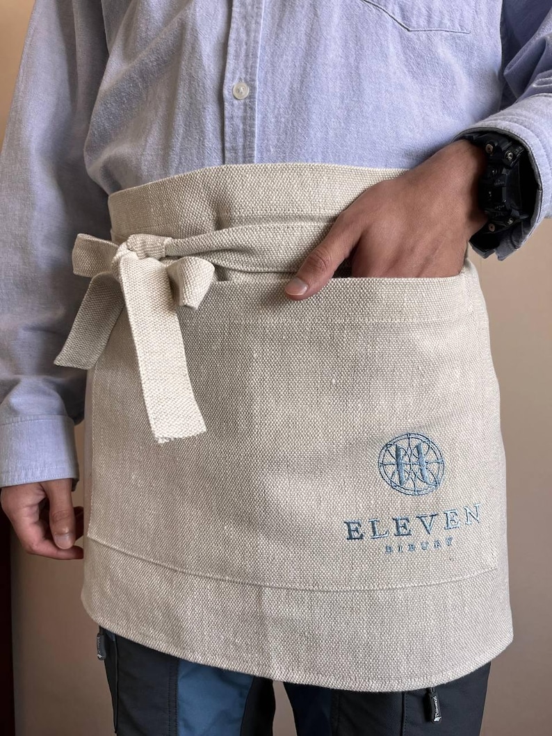 May include: A beige linen apron with a large pocket and a tie. The apron has a blue embroidered logo that says "ELEVEN" and "RESTAURANT".