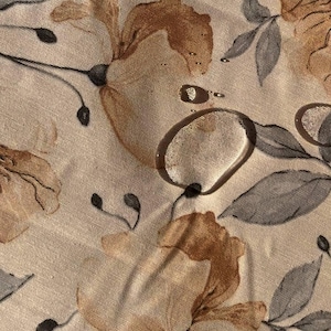 Puede incluir: Una tela beige con un patrón floral. Las flores son marrones y grises y tienen un efecto acuarela. La tela tiene una textura sutil.