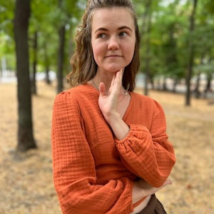 May include: An orange long-sleeved top with a V-neck and gathered sleeves. The top is paired with a brown skirt. The fabric appears to have a textured, crinkled appearance. The background is a blurred outdoor scene with trees.