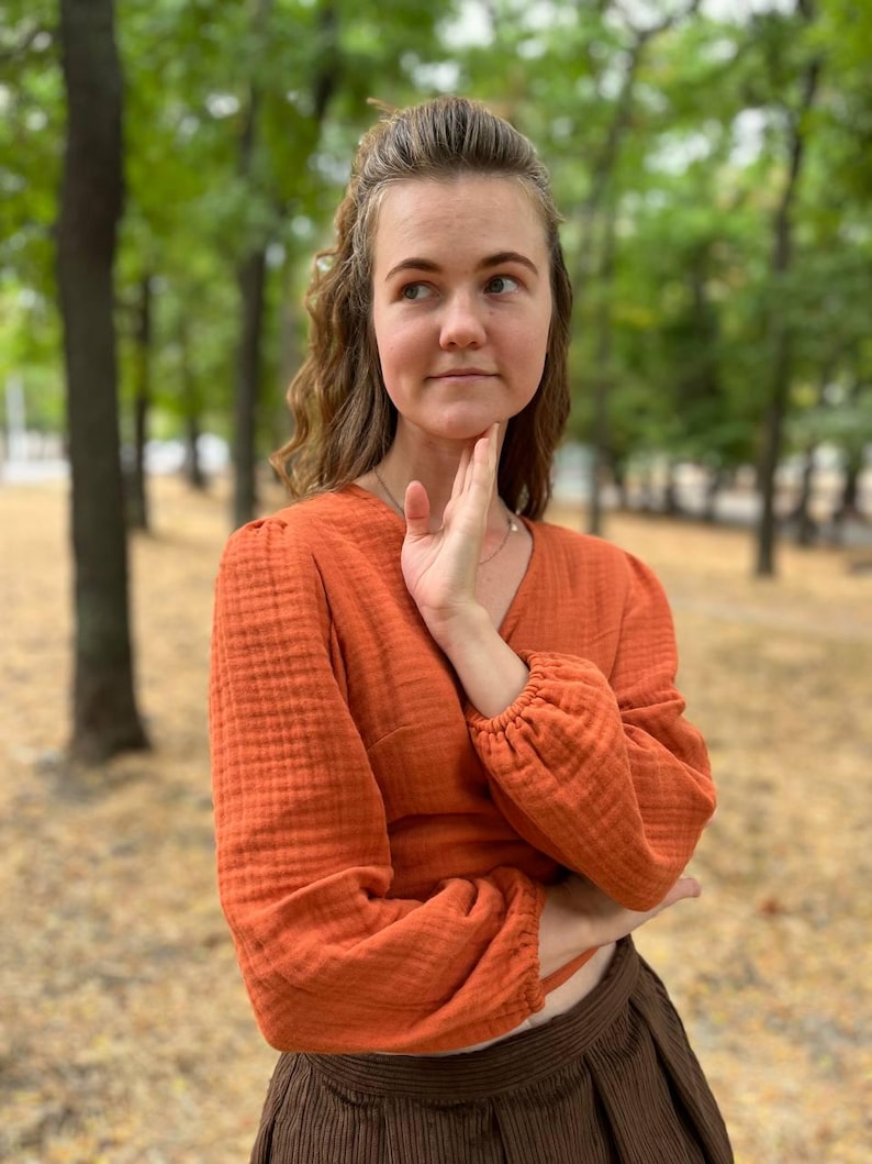 May include: An orange long-sleeved top with a V-neck and gathered sleeves. The top is paired with a brown skirt. The fabric appears to have a textured, crinkled appearance. The background is a blurred outdoor scene with trees.