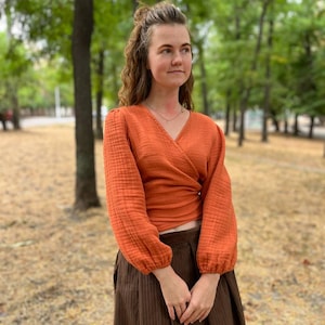 Puede incluir: Un top envolvente naranja con mangas largas y cuello en V. El top se combina con una falda marrón. Las mangas tienen un puño fruncido. El atuendo se usa al aire libre en un parque.