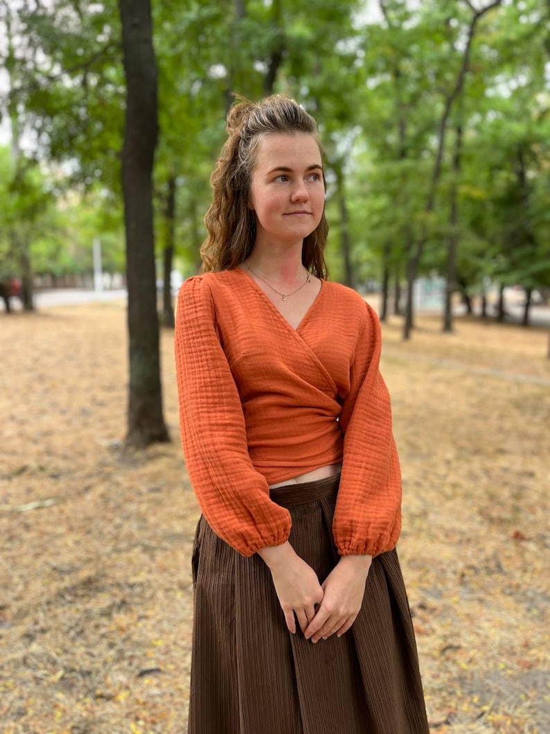 May include: An orange wrap top with long sleeves and a V-neck. The top is paired with a brown skirt. The sleeves have a gathered cuff. The outfit is worn outdoors in a park setting.