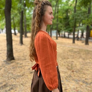 May include: An orange long-sleeve top with a wrap tie in the back, paired with a brown skirt. The top has a textured fabric and the skirt has vertical lines. The outfit is worn outdoors with a blurred background of trees.