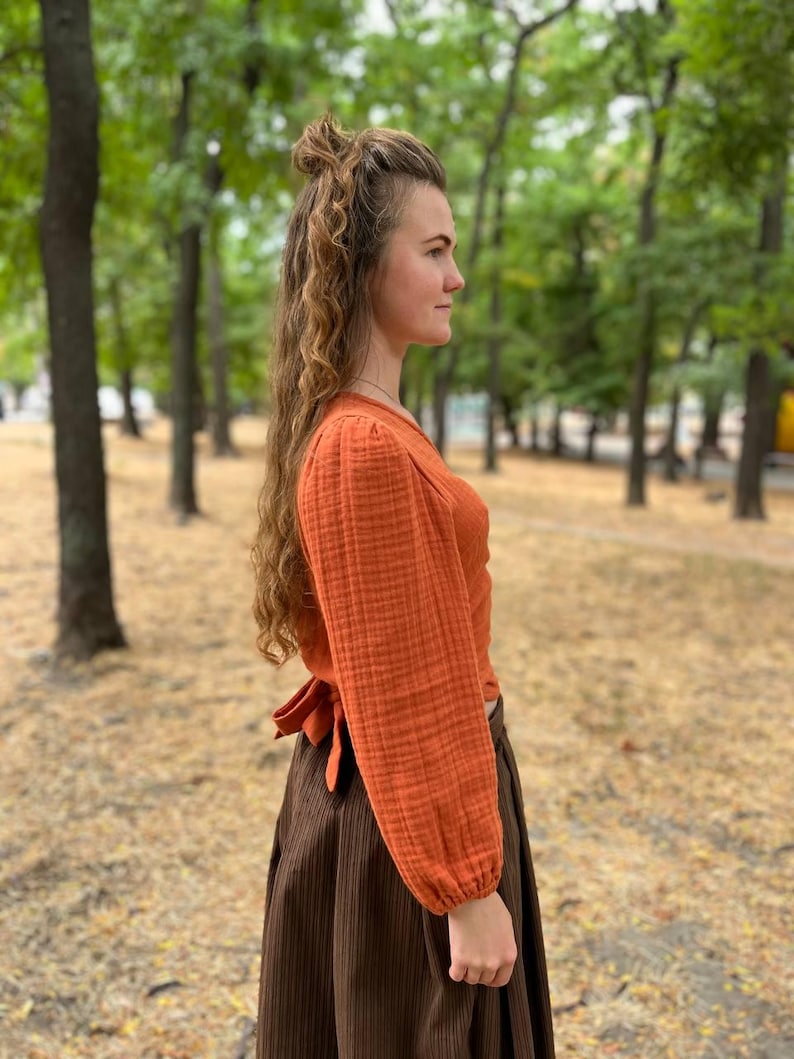 May include: An orange long-sleeve top with a wrap tie in the back, paired with a brown skirt. The top has a textured fabric and the skirt has vertical lines. The outfit is worn outdoors with a blurred background of trees.