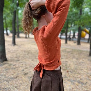 May include: An orange long-sleeved top with a tie at the waist and a brown skirt. The top has a textured appearance and the skirt has vertical stripes. The outfit is worn outdoors with a blurred green background.