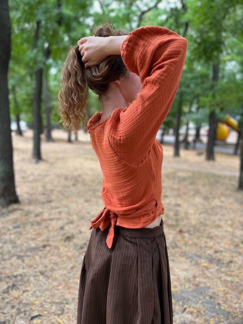 May include: An orange long-sleeved top with a tie at the waist and a brown skirt. The top has a textured appearance and the skirt has vertical stripes. The outfit is worn outdoors with a blurred green background.