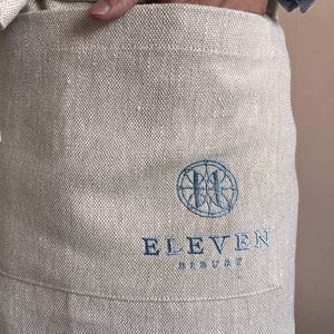 May include: A beige linen apron with a blue embroidered logo that says "ELEVEN BREWERY" and a circular design with a stylized "H" inside.