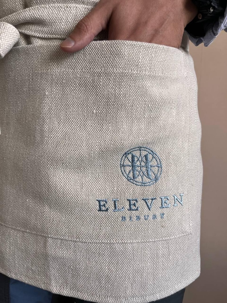 May include: A beige linen apron with a blue embroidered logo that says "ELEVEN BREWERY" and a circular design with a stylized "H" inside.