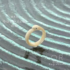 May include: A cream-coloured ring shaped like a snake, with a small black dot for an eye. The ring is set against a textured, teal-coloured background. Japanese text is visible in the background.