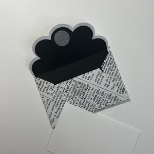 May include: A black and white envelope with a unique scalloped top, holding a white card. The envelope is printed with a newspaper-style design. The card is rectangular and blank.