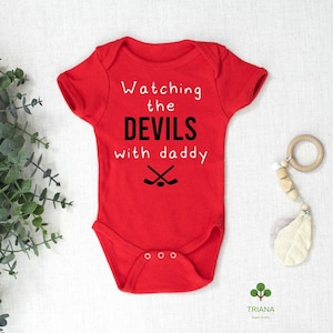 May include: Red baby bodysuit with the text "Watching the DEVILS with daddy" in white and black. Two crossed hockey sticks are below the text. The bodysuit has short sleeves and three snaps at the bottom.