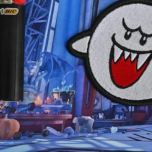 May include: A white and red Boo ghost character from the Super Mario Bros. video game series. The character is a plush patch with a stitched design. The patch is on a dark blue background with a chandelier and other decorations.