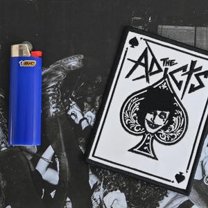 May include: White patch with black embroidery of a playing card with a spade suit and the text "The Adicts".