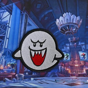 Puede incluir: Un fantasma blanco con una boca roja y dientes afilados, un personaje popular de videojuego, est&aacute; bordado en un parche de tela.