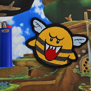 Puede incluir: Un personaje amarillo y negro parecido a una abeja con una boca roja y dientes afilados. El personaje est&aacute; bordado en un parche de tela.