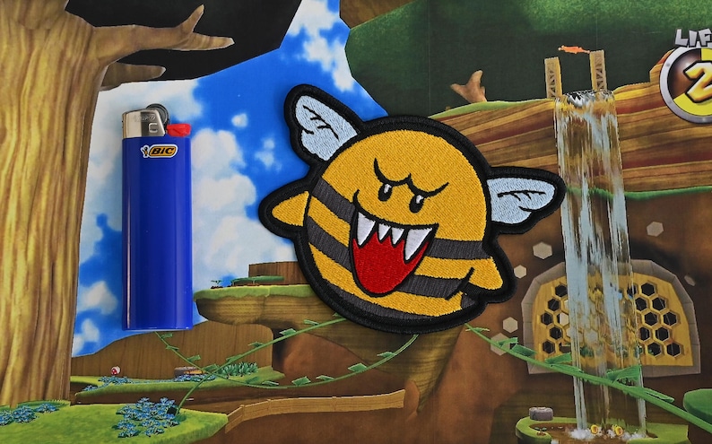 Puede incluir: Un personaje amarillo y negro parecido a una abeja con una boca roja y dientes afilados. El personaje est&aacute; bordado en un parche de tela.